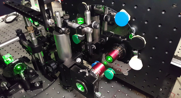 Optischer Aufbau mit verschiedensten diskreten Komponenten (Linsen, Spiegel etc.) auf einem schwingungsgedämpften optischen Labortisch, zur Vermessung eines Wafers. Grüner Schein eines Lasers in den optischen Komponenten.