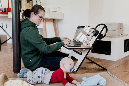 Frau arbeitet am Laptop am Boden in einer Wohnung sitzend, neben ihr krabbelt ein Baby. Foto: Standsome Worklifestyle